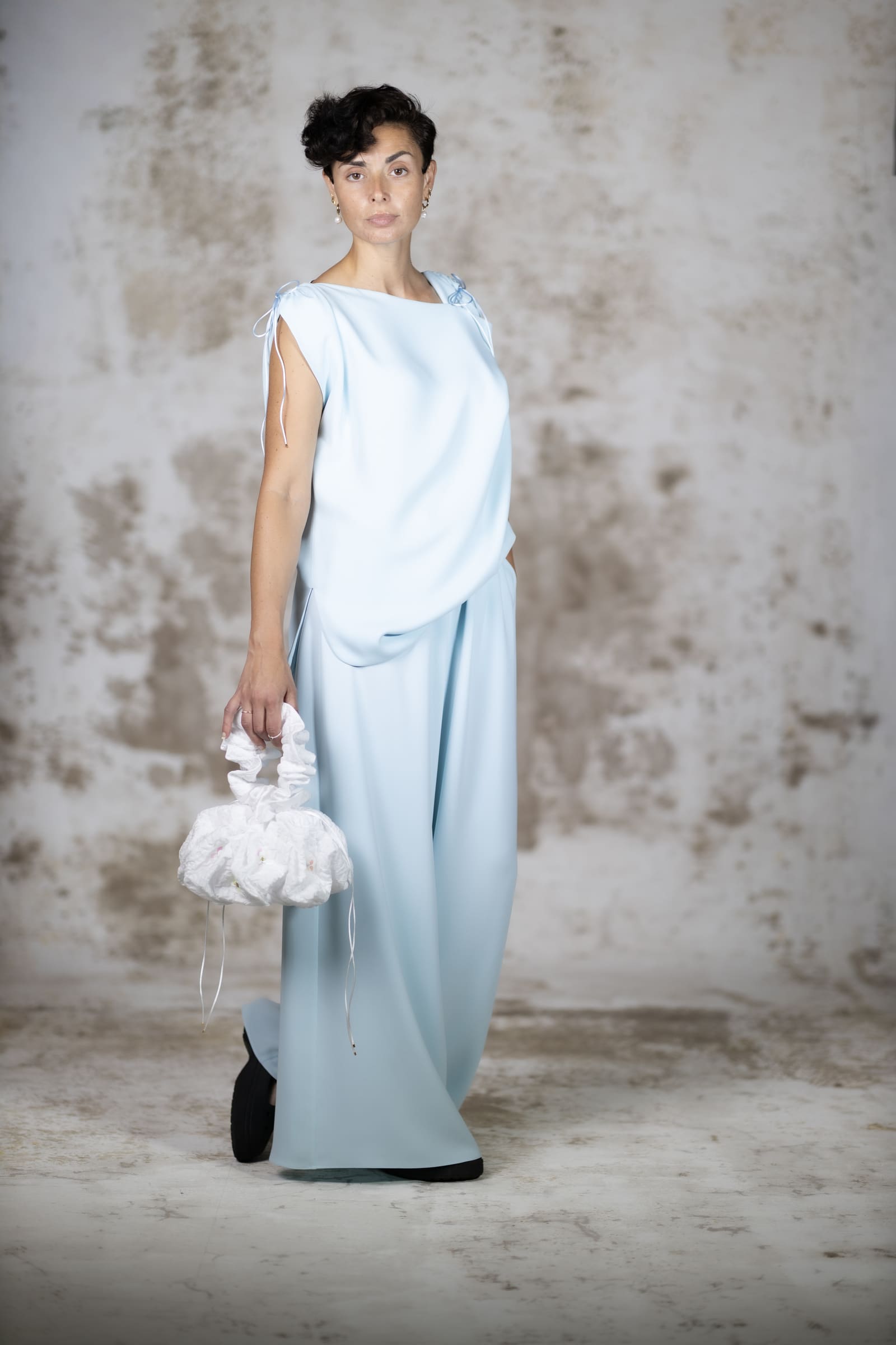 Full-body shot of a model wearing the Blue Pedra blouse, a loose minimalist light blue crepe top with a fluid draped silhouette and elegant ties - Anabel Santos