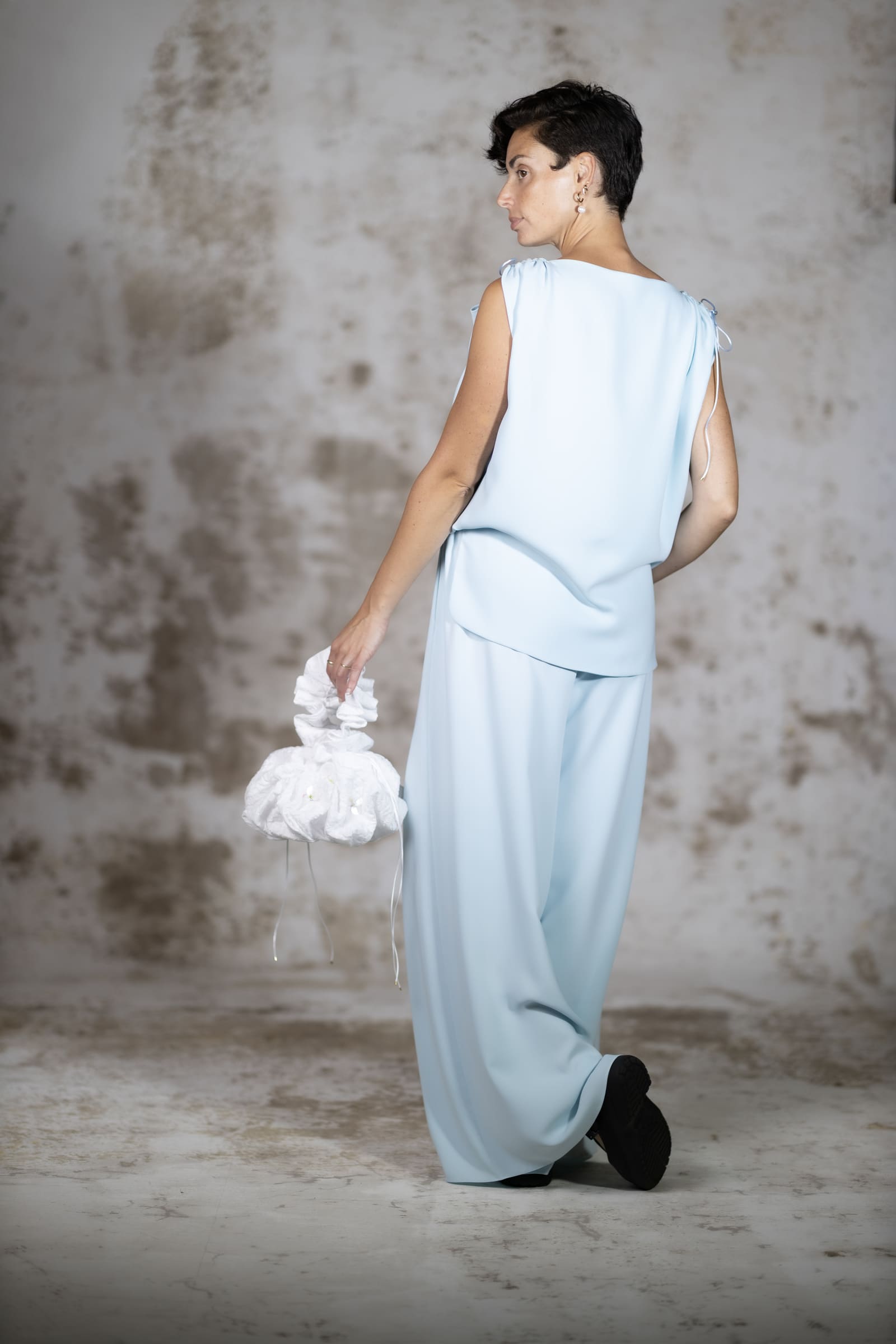 Full-body back view of the Blue Pedra blouse, showing the elegant clean drape of the minimalist light blue crepe fabric and handcrafted rat-tail tie details - Anabel Santos